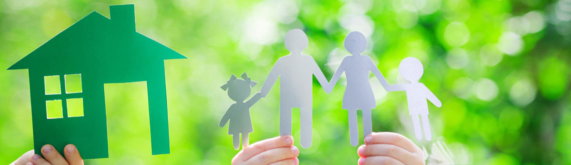 Papierausschnitte eines Hauses und einer Familie, die von Händen vor einem grünen, verschwommenen Hintergrund gehalten werden.